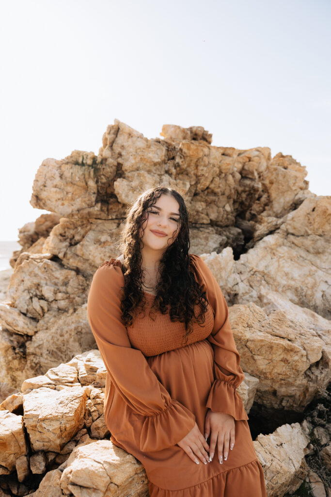 Maleah looking at the camera while sitting on a rock posing with both hands on her thighs.